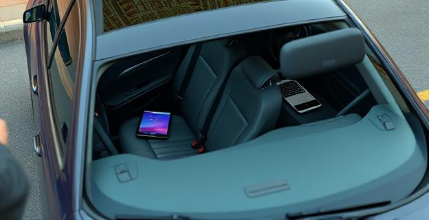 Un coche aparcado en la calle con una tablet y un portátil dentro, ambos ocultos bajo los asientos. Un ladrón con un dispositivo móvil en mano buscando señales inalámbricas. Detectores de baterías de litio junto al ladrón.