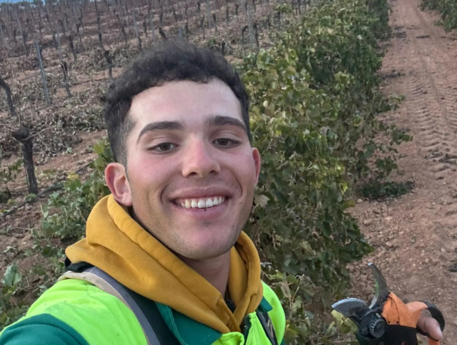 Un paisaje agrícola con cultivos leñosos como viñas, almendros y olivos bajo un cielo despejado. En el centro, un joven agricultor con herramientas de trabajo en un campo de Castilla-La Mancha.